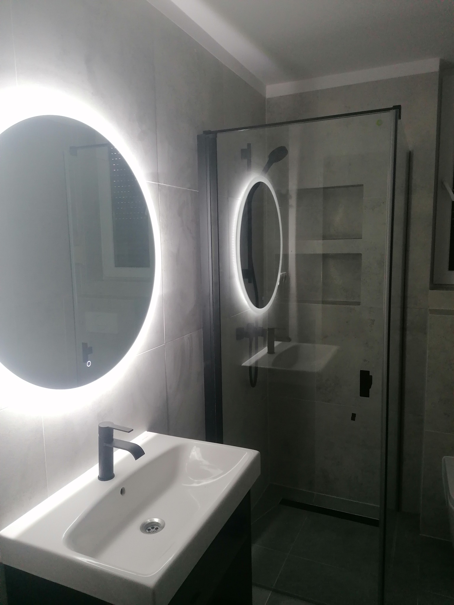 Nowoczesna łazienka z szarymi płytkami, okrągłym lustrem LED i kabiną prysznicową z czarnymi profilami, prezentująca minimalistyczny design i funkcjonalne rozwiązania.