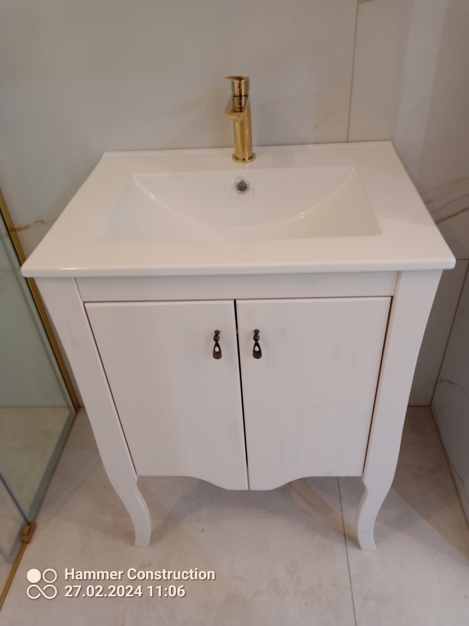 Biała szafka pod umywalkę z ozdobnymi nóżkami i złotą baterią, widoczna w łazience z marmurowymi płytkami.