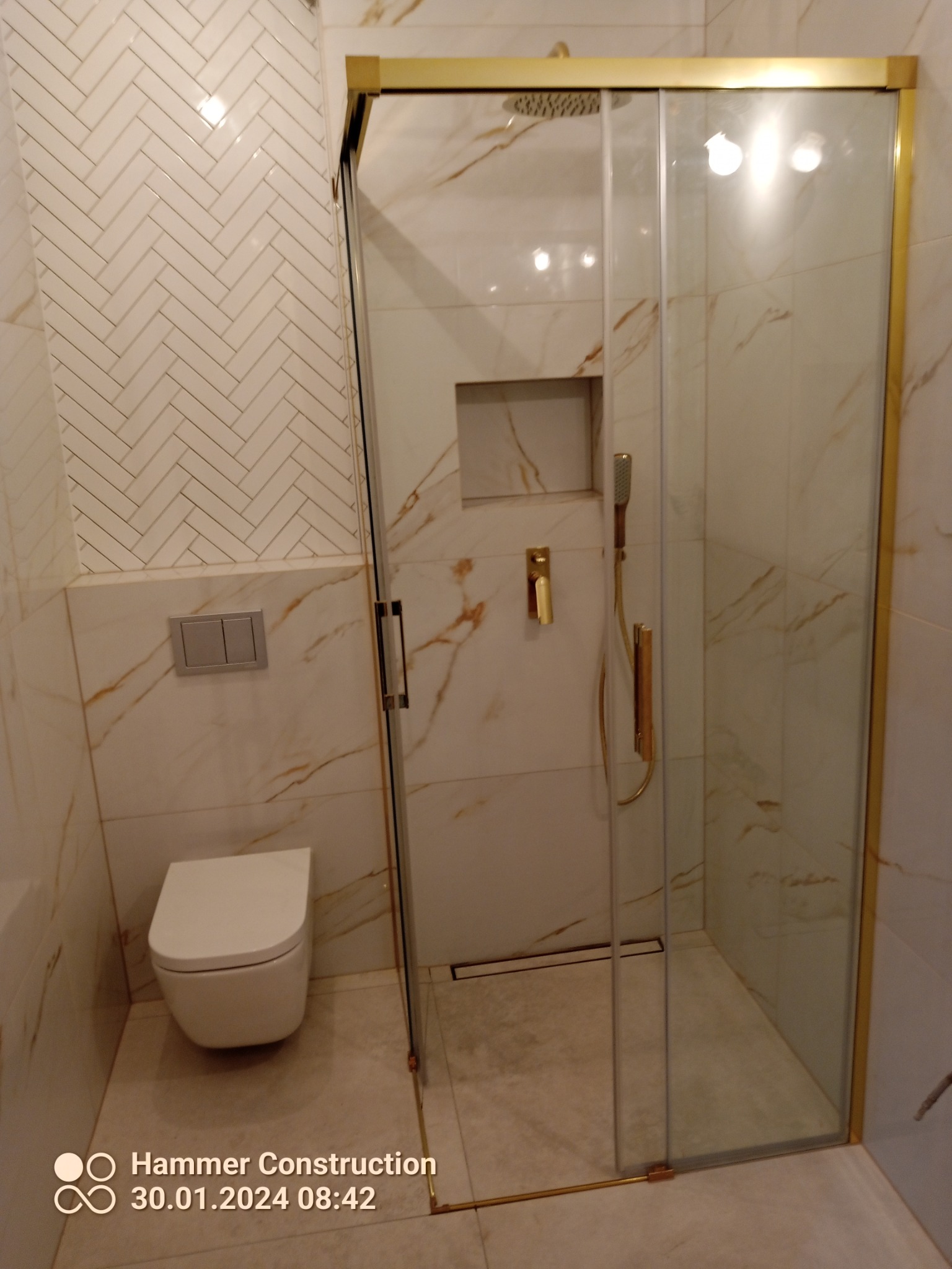 Nowoczesna łazienka z kabiną prysznicową w złotej ramie, białą ceramiką i marmurkowymi płytkami ułożonymi w jodełkę.