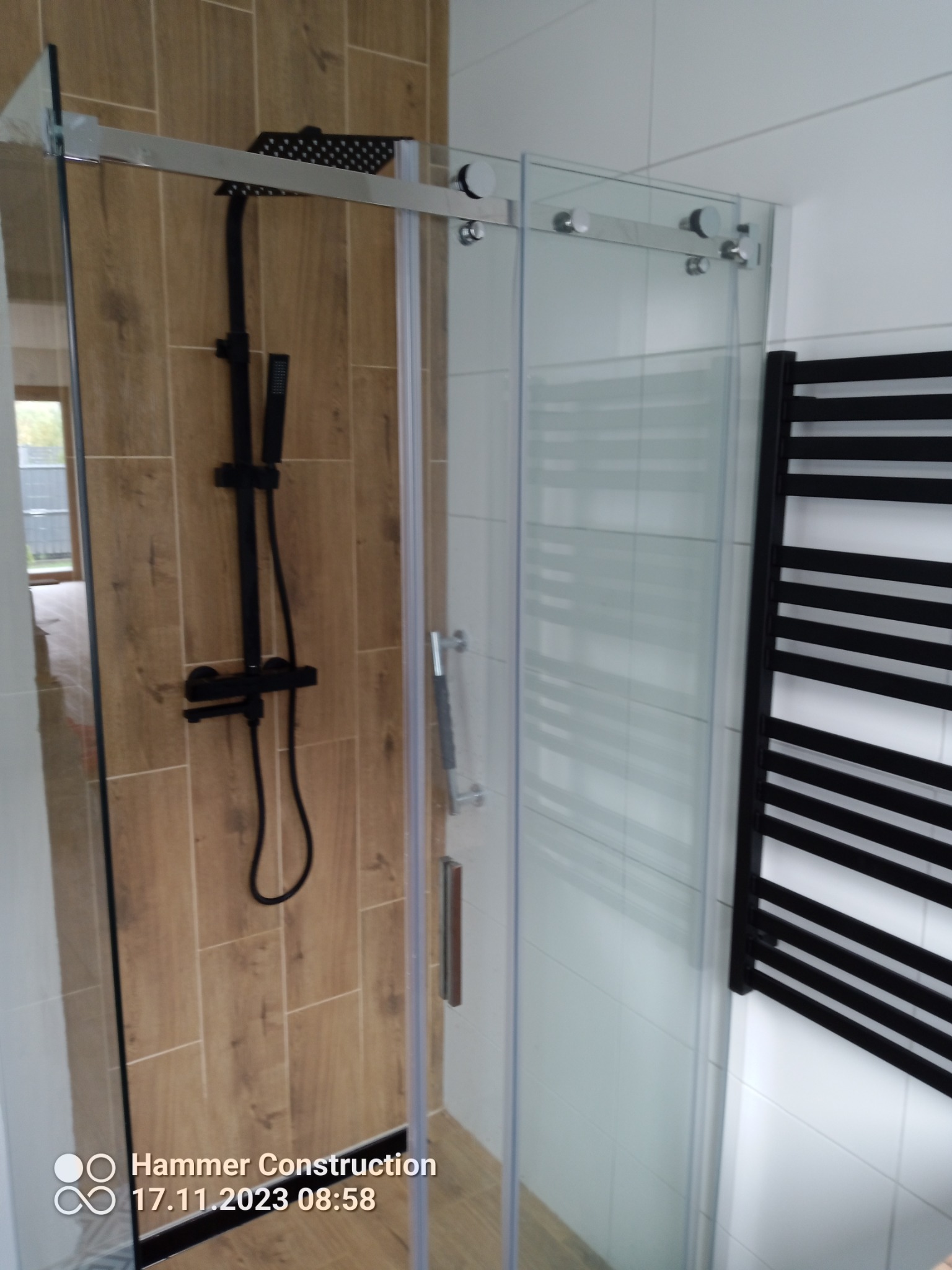 Kabina prysznicowa z przesuwnymi drzwiami, czarna bateria i deszczownica na ścianie wyłożonej płytkami imitującymi drewno, obok grzejnik drabinkowy w kolorze czarnym.