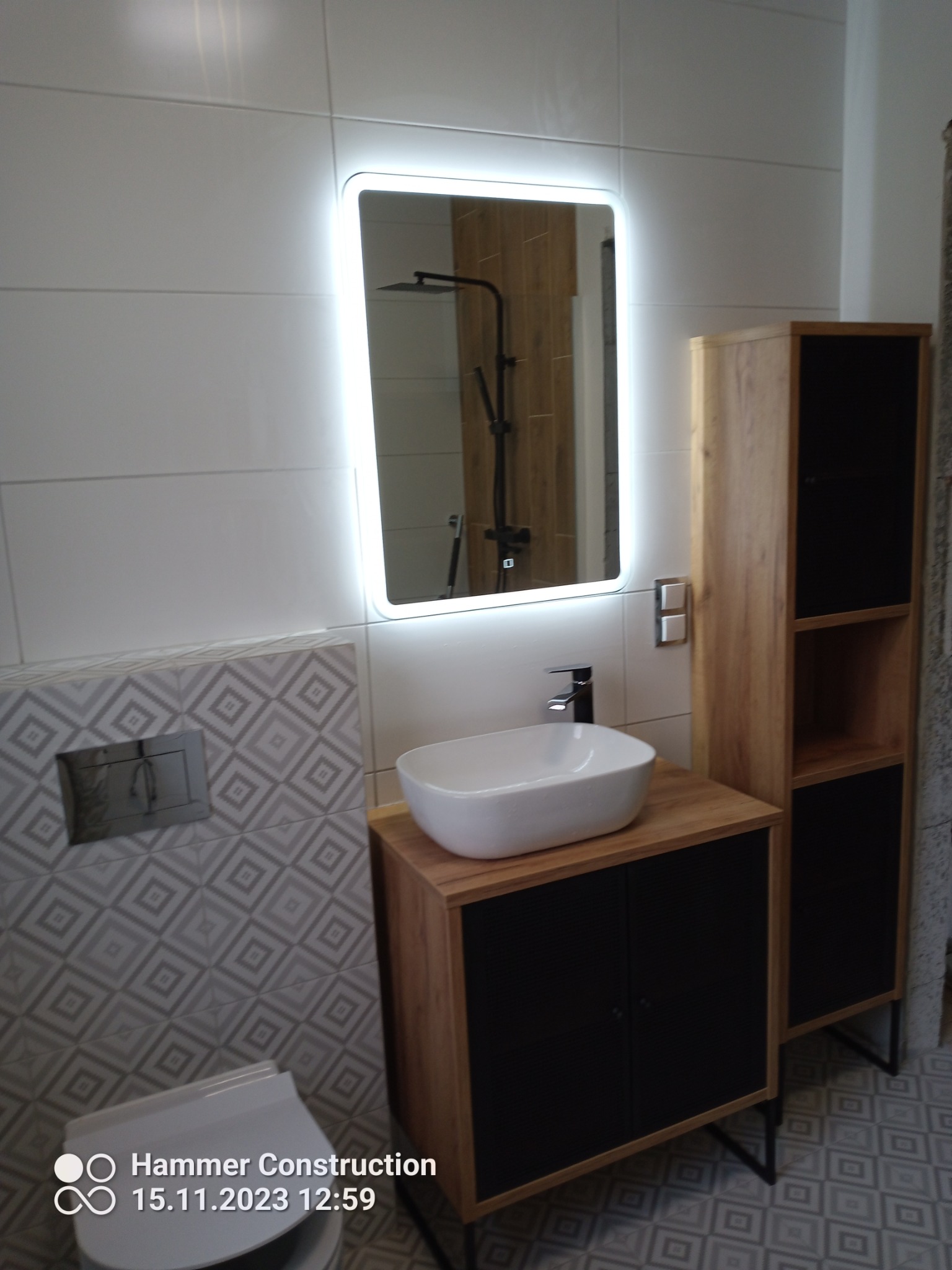 Nowoczesna łazienka z podświetlanym lustrem, umywalką nablatową na szafce w kolorze drewna z czarnymi frontami, oraz wysoką szafką w podobnej stylistyce, na tle płytek w geometryczny wzór.