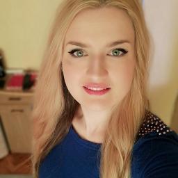 Projekt Zdrowie Nowy Sącz - Portret uśmiechniętej blondynki w niebieskiej bluzce z ozdobnymi aplikacjami na ramionach, z makijażem oczu, na tle jasnej ściany i mebli.