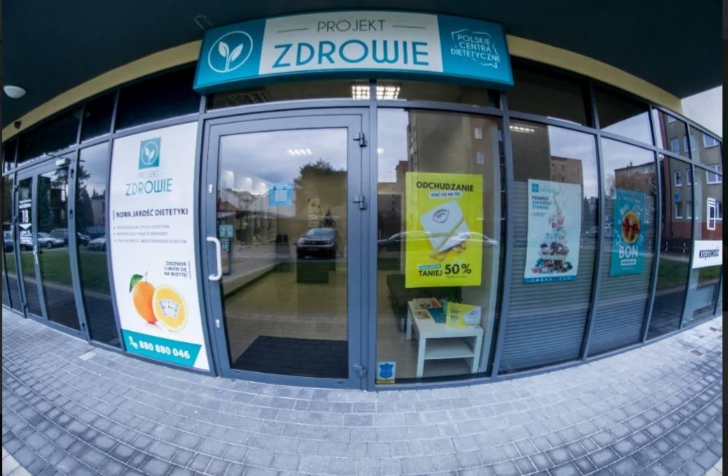 Fasada budynku z dużymi oknami, szyld 'Projekt Zdrowie - Polskie Centra Dietetyczne' nad wejściem, plakaty o odchudzaniu i promocjach w oknach.