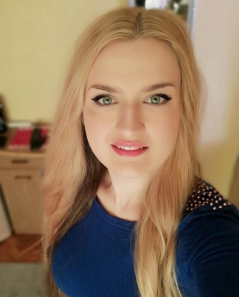 Portret uśmiechniętej blondynki w niebieskiej bluzce z ozdobnymi aplikacjami na ramionach, z makijażem oczu, na tle jasnej ściany i mebli.