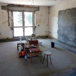 Pokój w trakcie remontu: odsłonięte fragmenty ścian, widoczne rury instalacji przy podłodze, przygotowane narzędzia i materiały budowlane, okno z widokiem na zieleń.