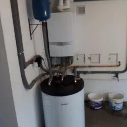 Biały piec gazowy Vaillant z niebieskim zbiornikiem wyrównawczym, szarymi rurami i dwoma otwartymi wiadrami z farbą na betonowej podłodze w pomieszczeniu gospodarczym.