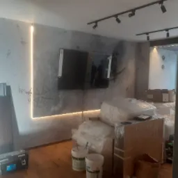 Pokój po remoncie z ozdobną ścianą o fakturze betonu, podświetloną pionowo LED, z widocznym kompasem na ścianie, narzędziami i materiałami budowlanymi na podłodze.