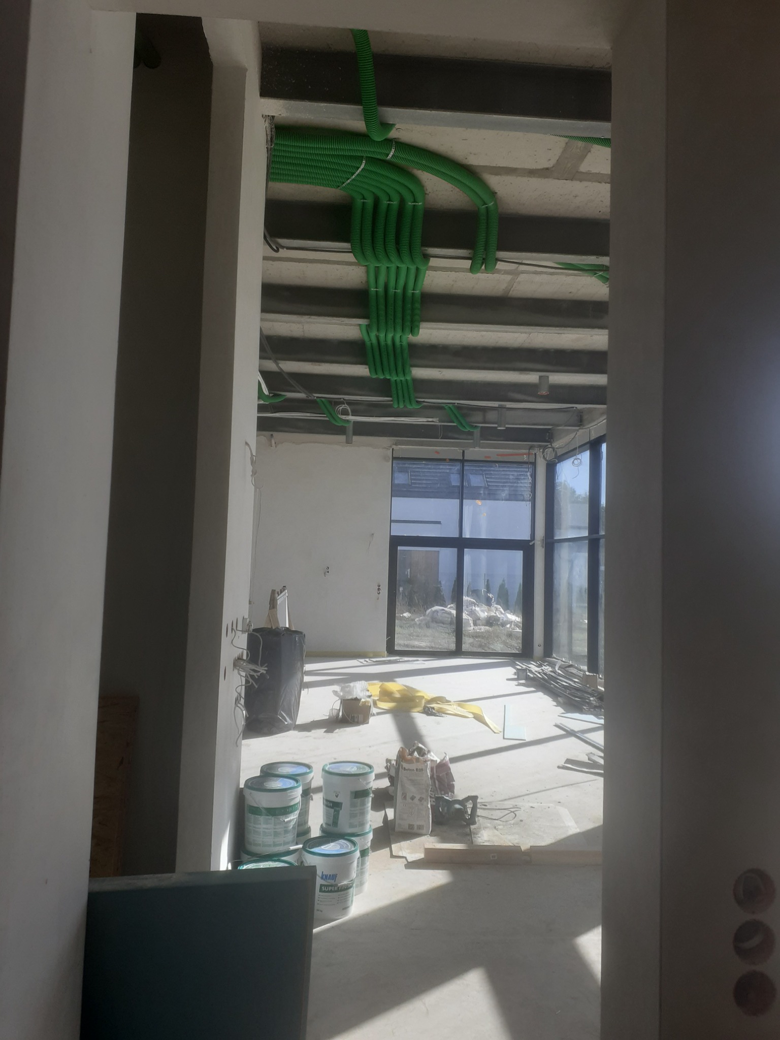 Wnętrze w trakcie remontu, widoczne puszki z masą szpachlową, worki z zaprawą, zielone peszle na suficie, duże okna i surowy stan ścian.