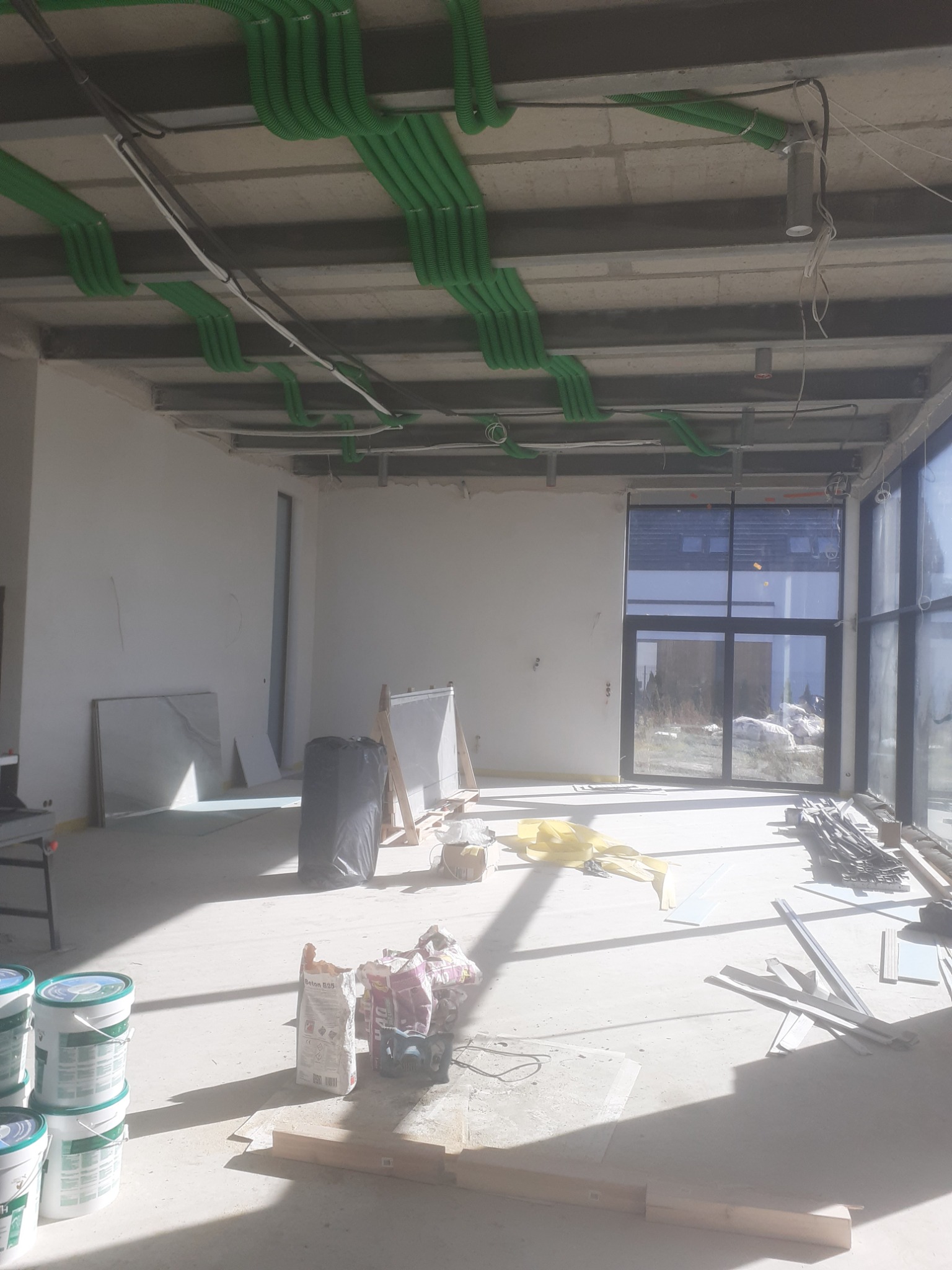 Wnętrze w trakcie wykończenia: widoczne zielone peszle na suficie, przygotowane płyty, worki z zaprawą i puste wiadra po farbie na jasnej podłodze, duże okno z widokiem na zewnątrz.