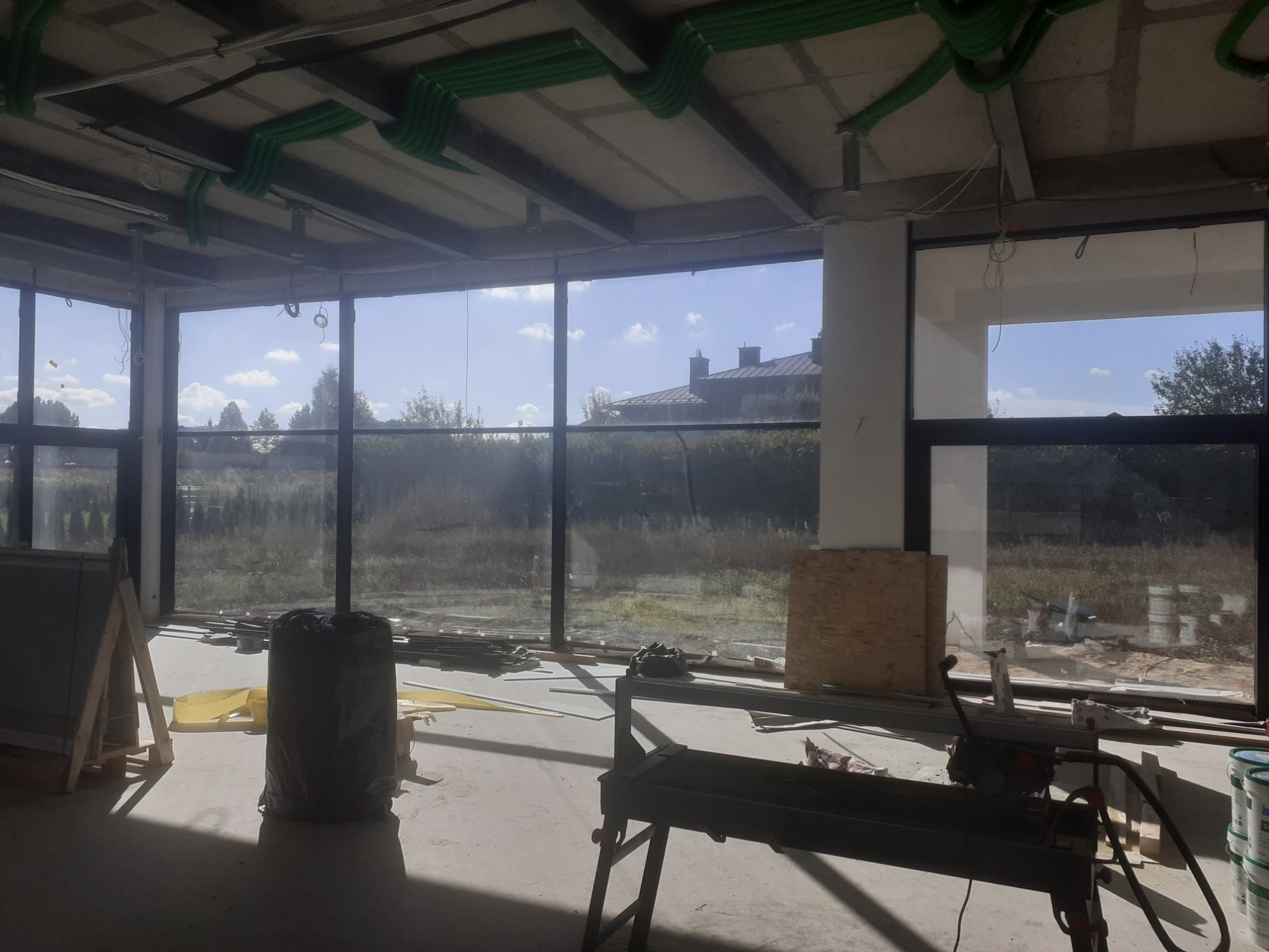 Wnętrze w trakcie remontu z widocznymi elementami instalacji pod sufitem, narzędziami do cięcia płytek, workiem z odpadami i dużymi oknami z widokiem na zewnątrz.