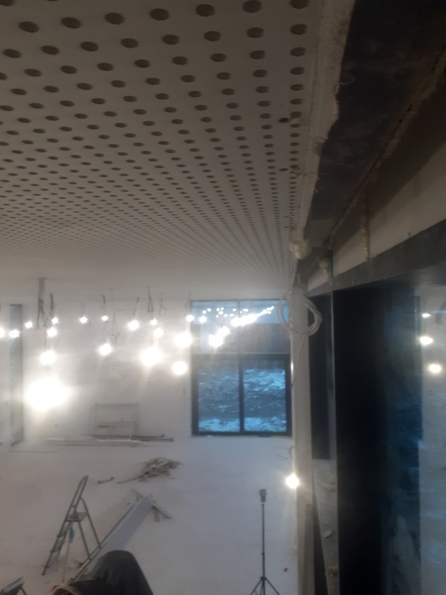 Montaż perforowanego sufitu podwieszanego z płyt kartonowo-gipsowych w jasnym pomieszczeniu z widocznymi przewodami, oświetleniem tymczasowym i drabiną.