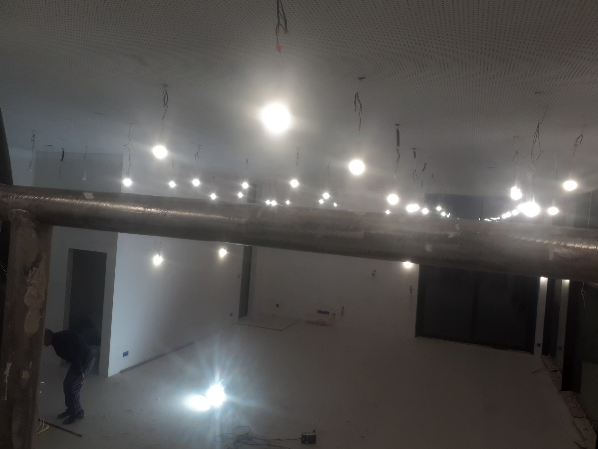 Wnętrze w trakcie prac wykończeniowych, widoczne surowe ściany, tymczasowe oświetlenie, pracownik w tle i metalowa rura na pierwszym planie.