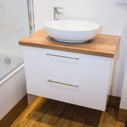Biała szafka łazienkowa z dwiema szufladami i blatem z drewna, na której stoi okrągła umywalka. W tle widoczna kabina prysznicowa i lustro.