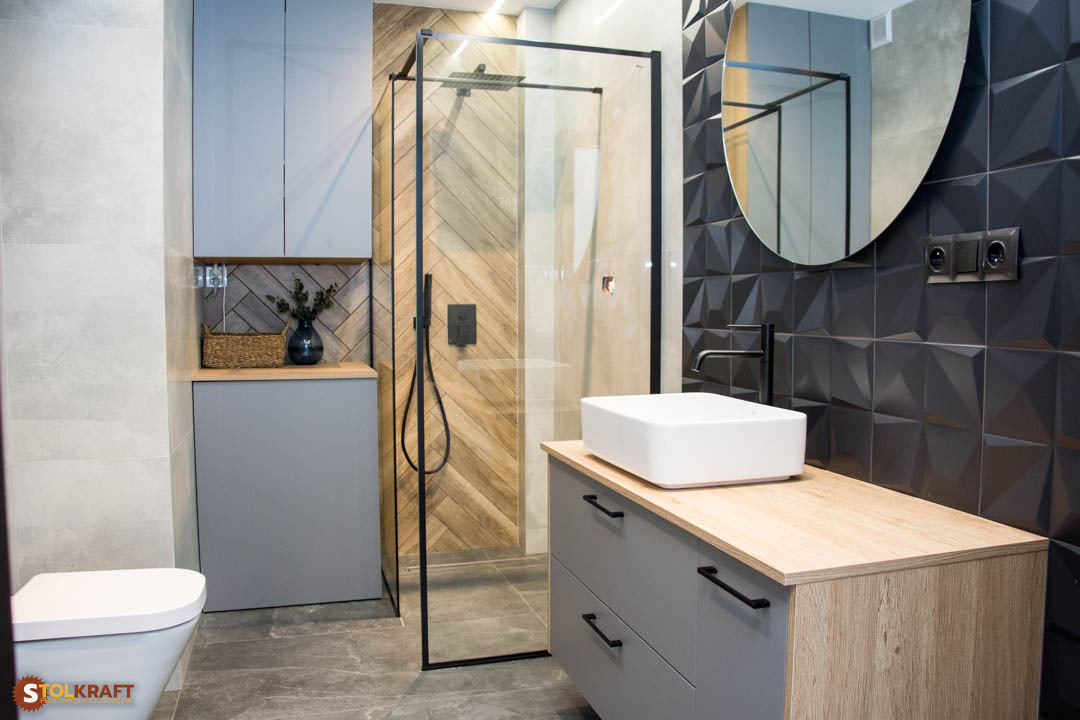 Nowoczesna łazienka z szarą zabudową meblową, drewnianym blatem, kabiną prysznicową ze szklaną ścianką i czarnymi detalami oraz umywalką nablatową.