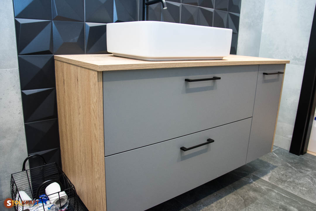 Szara szafka łazienkowa z drewnianymi bokami i blatem, biała umywalka nablatowa, czarne uchwyty i trójwymiarowe czarne płytki na ścianie w tle.