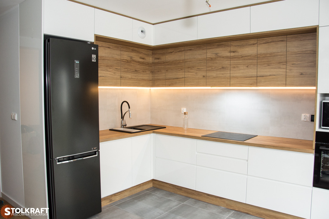 Nowoczesna kuchnia w kształcie litery L z białymi szafkami dolnymi i drewnianymi szafkami górnymi, podświetlona listwą LED, z czarną lodówką i szarą podłogą.