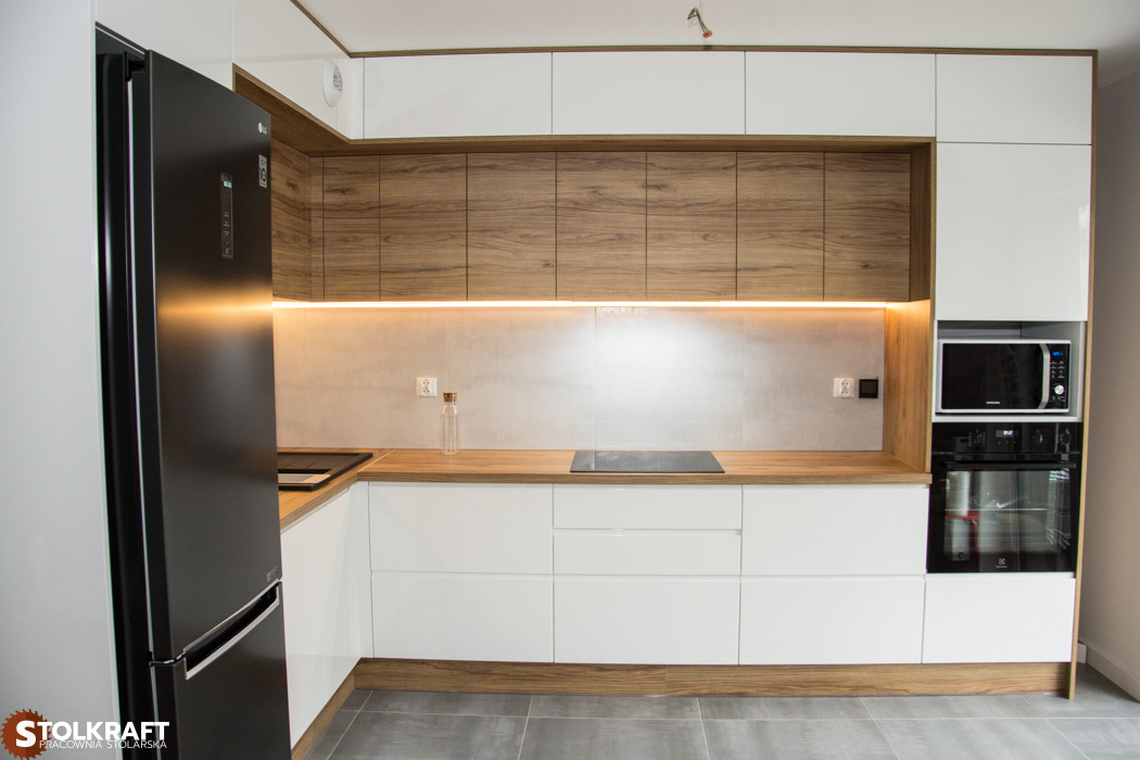 Nowoczesna kuchnia na wymiar z białymi frontami, drewnianymi akcentami i oświetleniem LED pod szafkami wiszącymi, widoczny czarny sprzęt AGD.