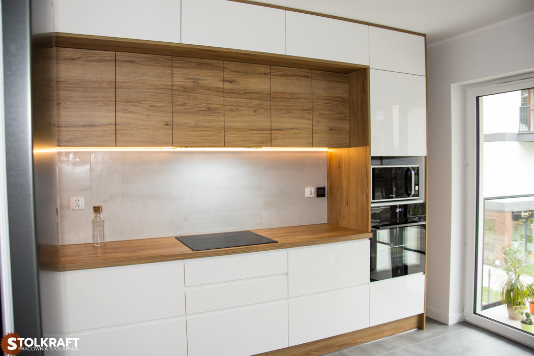 Nowoczesna kuchnia: białe lakierowane fronty dolne, drewniane szafki górne z podświetleniem LED, drewniany blat, płyta indukcyjna, piekarnik i mikrofalówka w zabudowie, widok na zewnątrz przez okno.