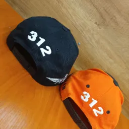 Dwie czapki typu snapback leżące na drewnianym blacie: czarna z białym haftem '312' i logo ze skrzydłami, oraz pomarańczowa z białym haftem '312'.