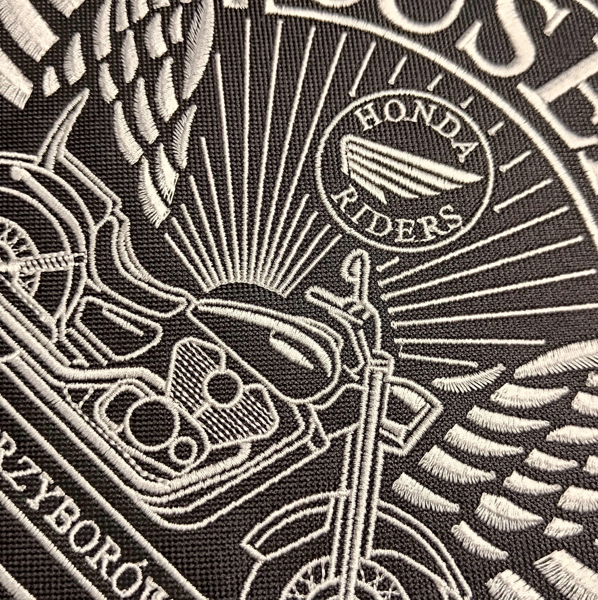 Czarno-biały haftowany emblemat z motywem motocykla Honda Riders, z elementami graficznymi imitującymi skrzydła i promienie słońca, na teksturowanym materiale.