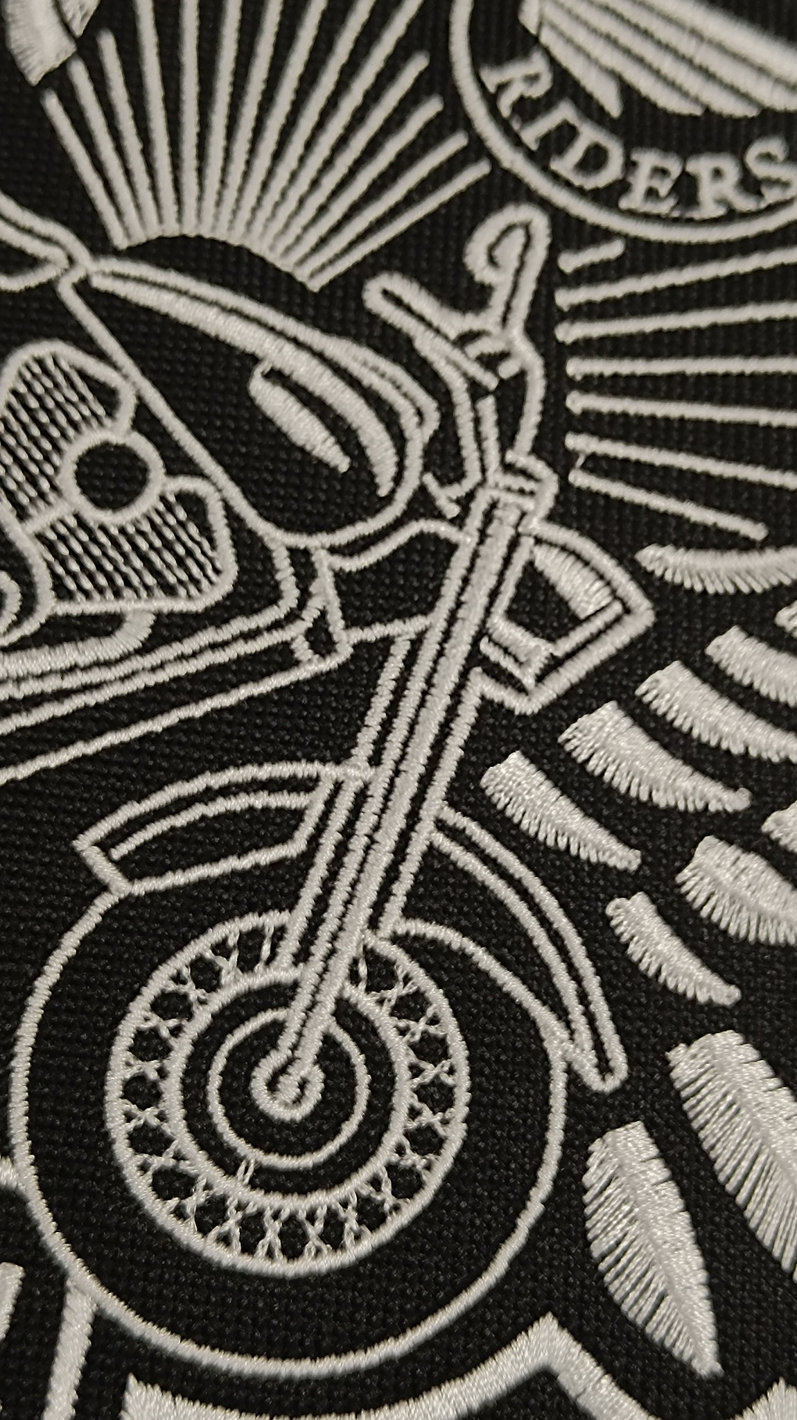 Czarno-biały detal haftu przedstawiający motocykl i napis 'RIDERS', z widoczną teksturą tkaniny.