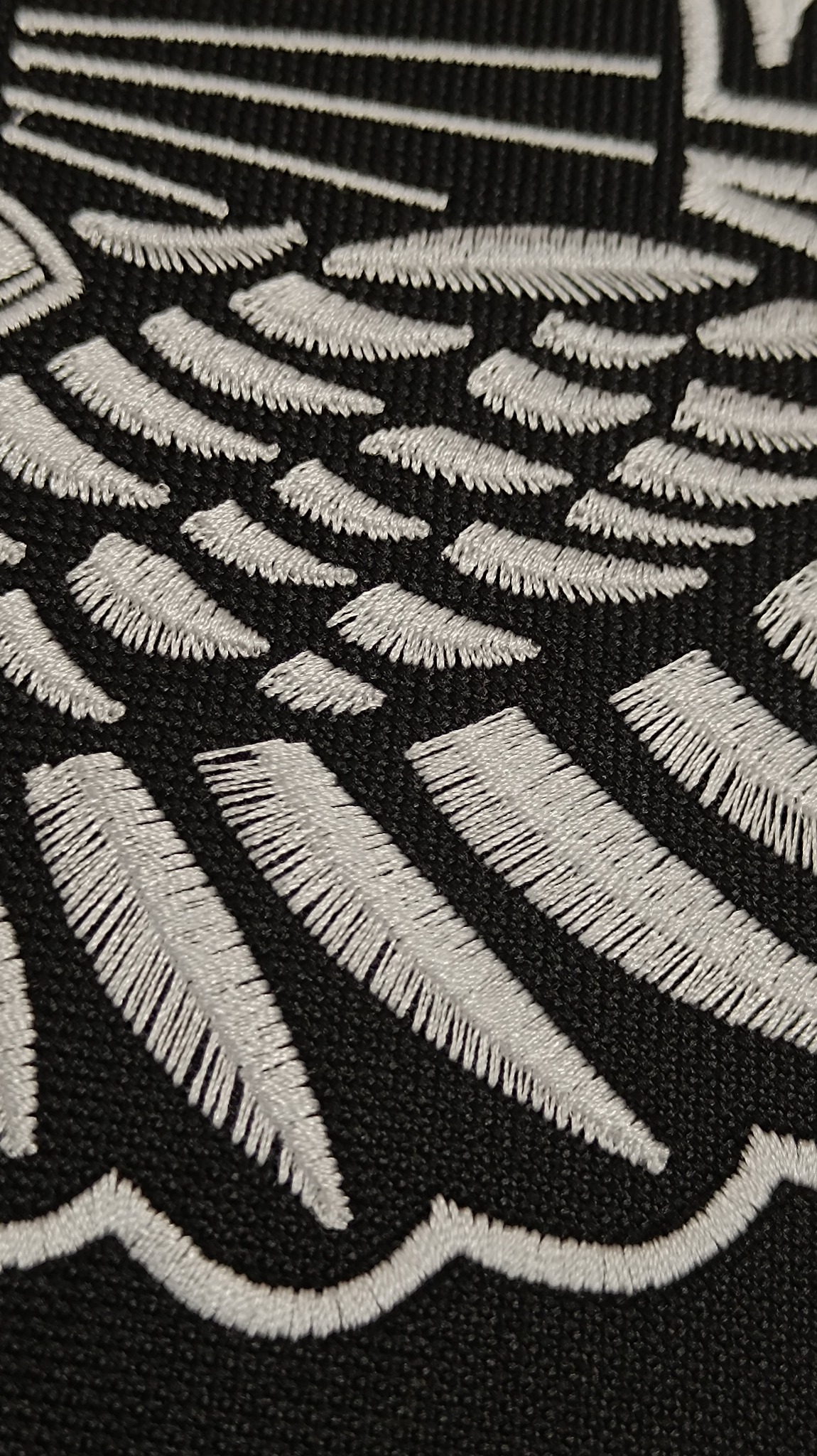 Zbliżenie na fragment czarnej tkaniny z białym, gęstym wzorem przypominającym pióra lub promienie, wykonanym techniką haftu.