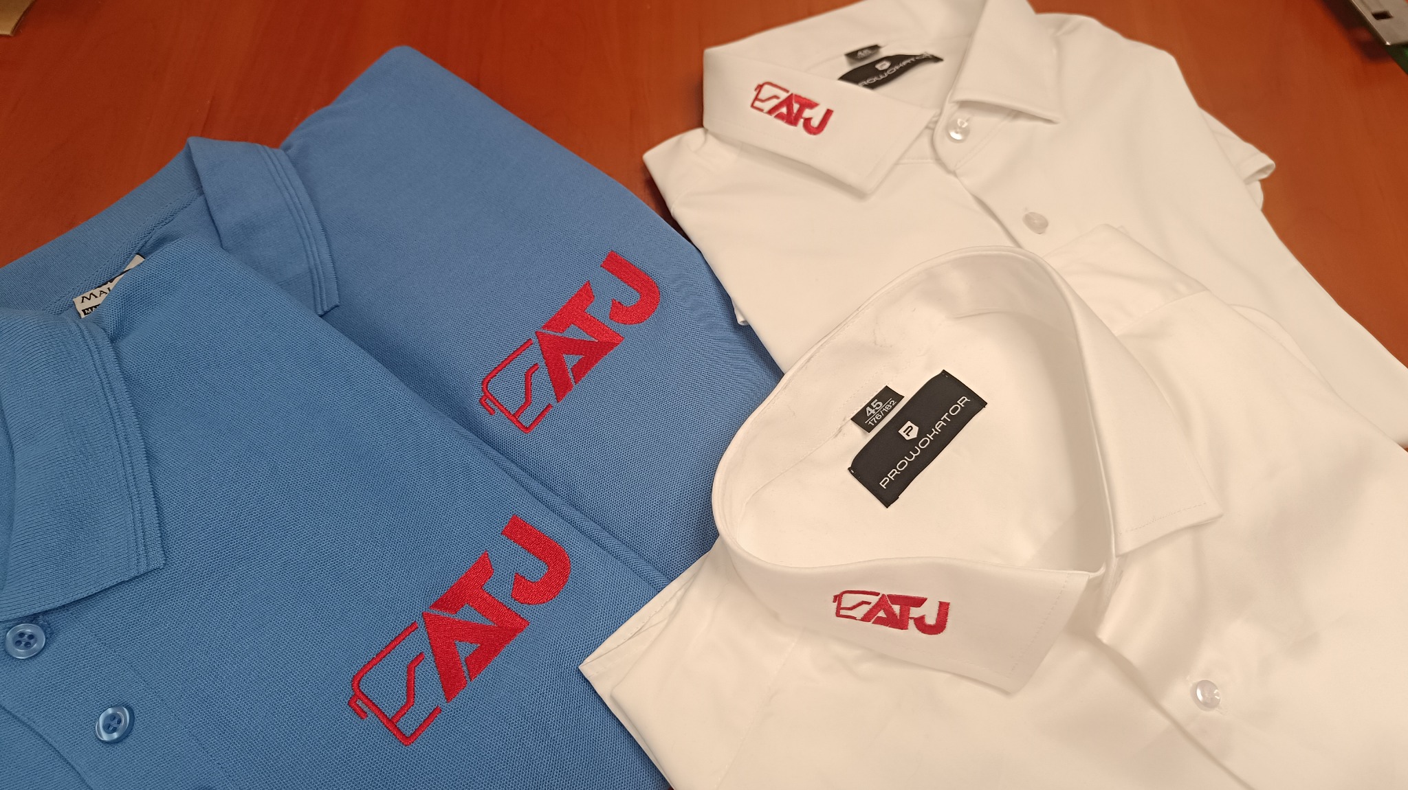 Dwa polo w kolorze błękitnym i dwie białe koszule z krótkim rękawem, wszystkie z wyhaftowanym czerwonym logo 'EATJ' na kołnierzyku lub na piersi.