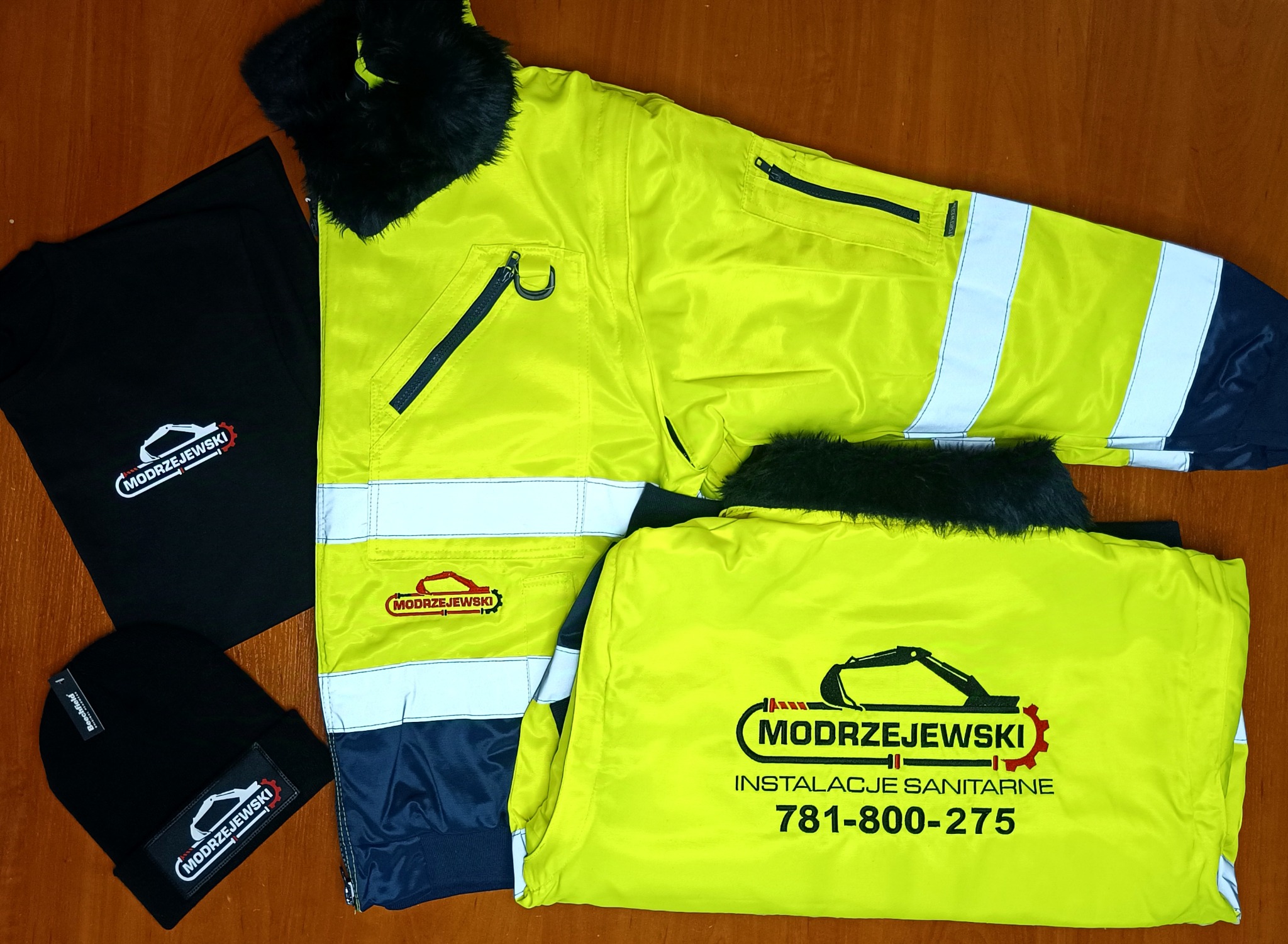 Żółta kurtka robocza z futrzanym kołnierzem, czarna koszulka i czapka z logo firmy instalacyjnej z motywem koparki, leżące na drewnianym blacie.