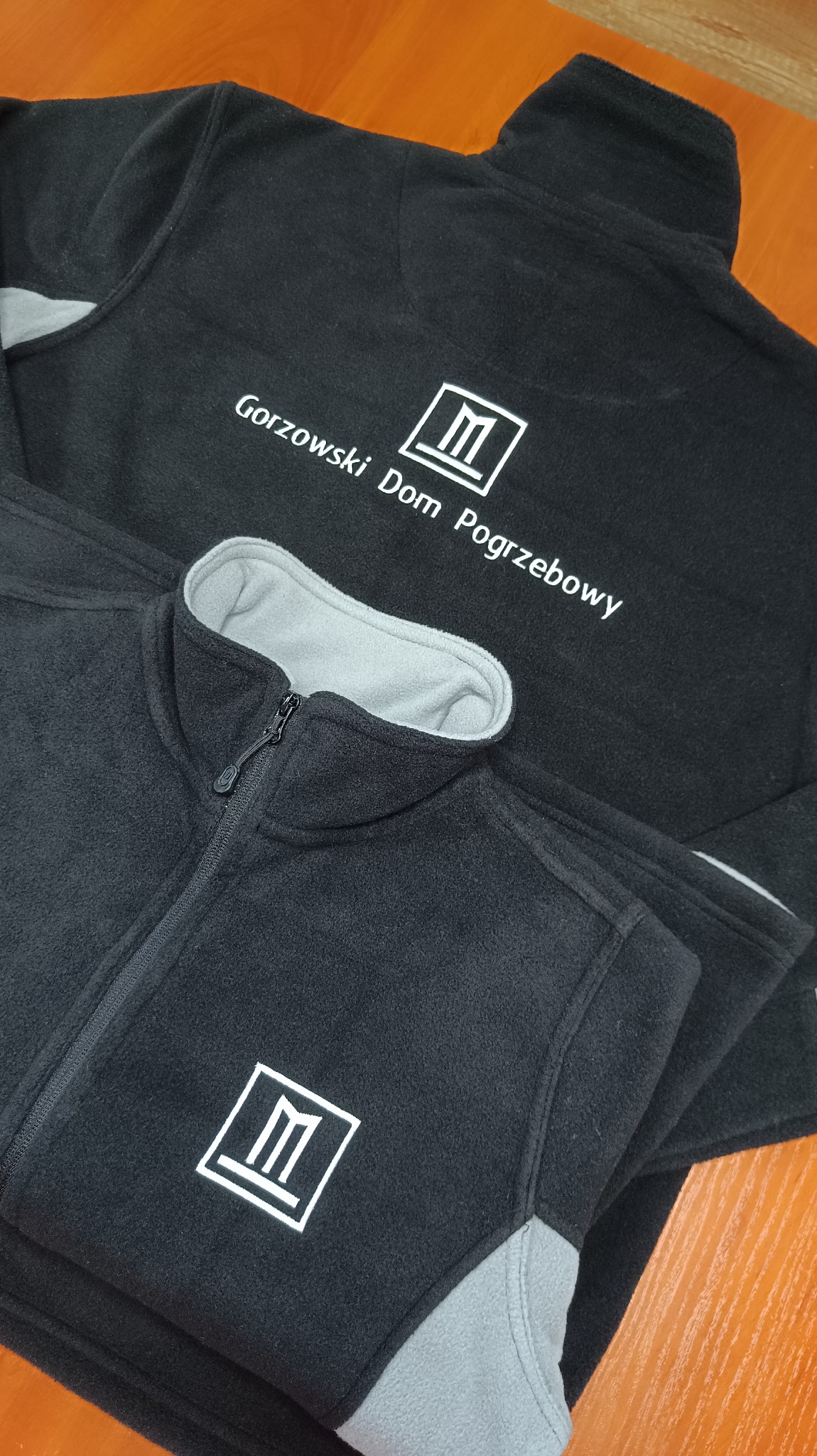 Dwie czarne bluzy polarowe z suwakami, na których widnieje białe logo i napis 'Gorzowski Dom Pogrzebowy'. Jedna bluza jest częściowo rozpięta.