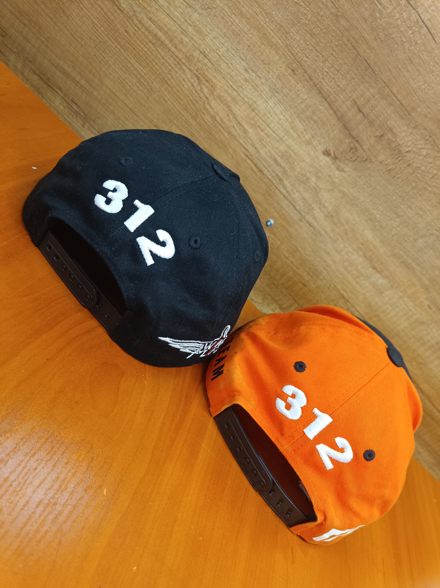Dwie czapki typu snapback leżące na drewnianym blacie: czarna z białym haftem '312' i logo ze skrzydłami, oraz pomarańczowa z białym haftem '312'.