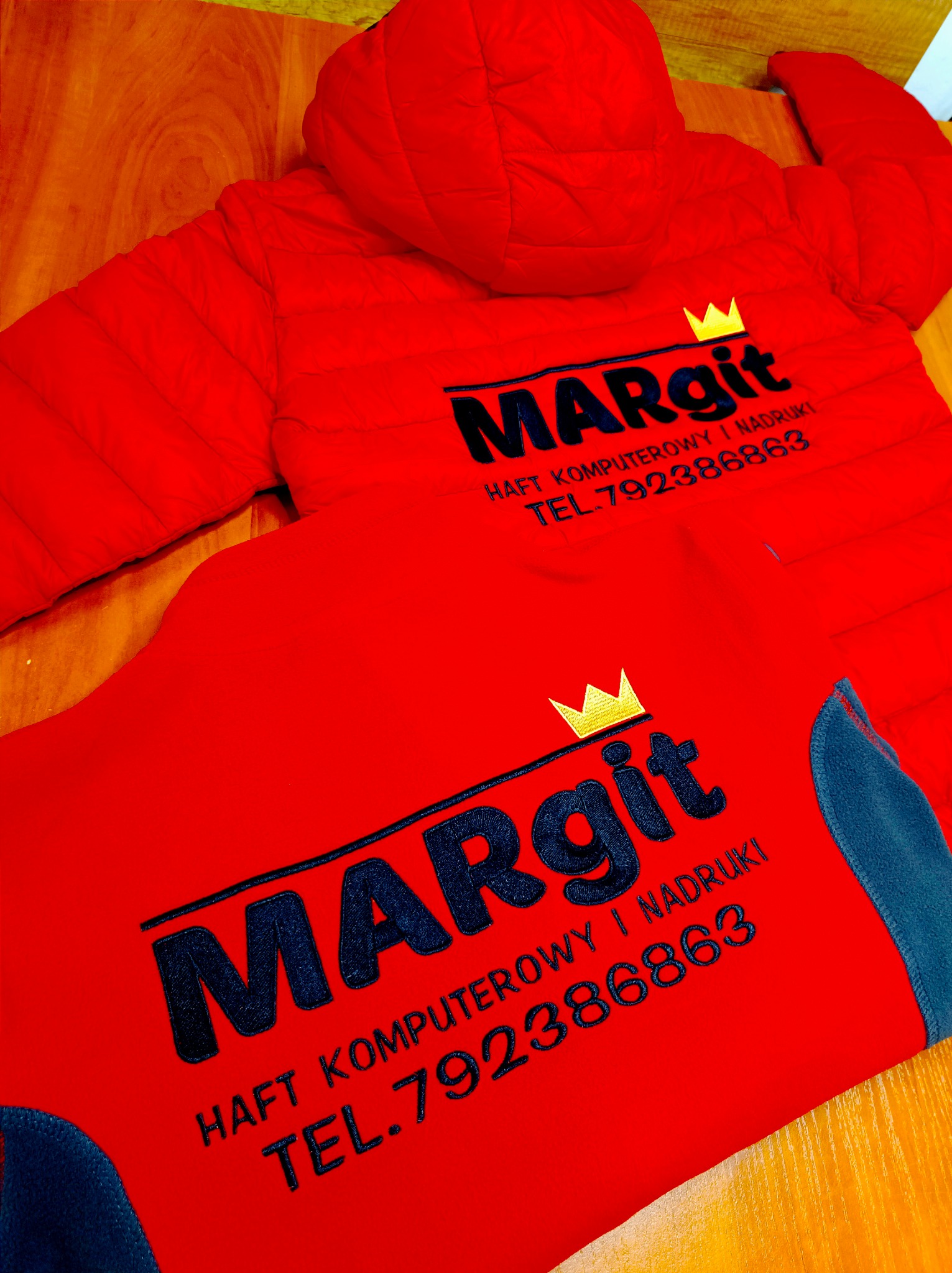 Czerwona kurtka i bluza z wyhaftowanym czarnym napisem 'Margit HAFT KOMPUTEROWY I NADRUKI TEL.792386863' oraz złotą koroną nad napisem.