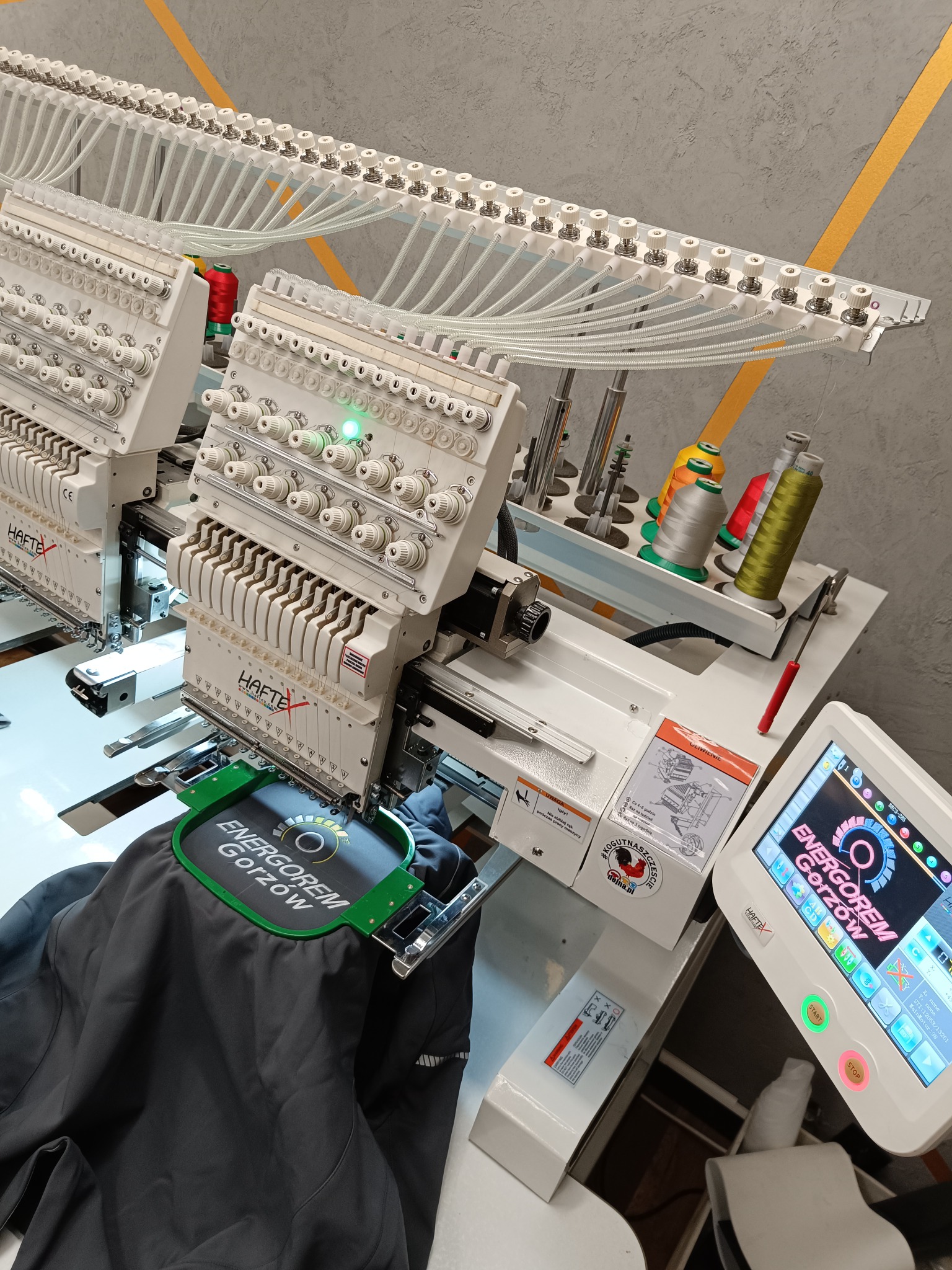 Wielogłowicowa maszyna hafciarska w trakcie nanoszenia logo 'ENERGOREM Gorzów' na ciemną kurtkę, widoczne szpulki z kolorowymi nićmi i panel sterowania z wyświetlanym wzorem.