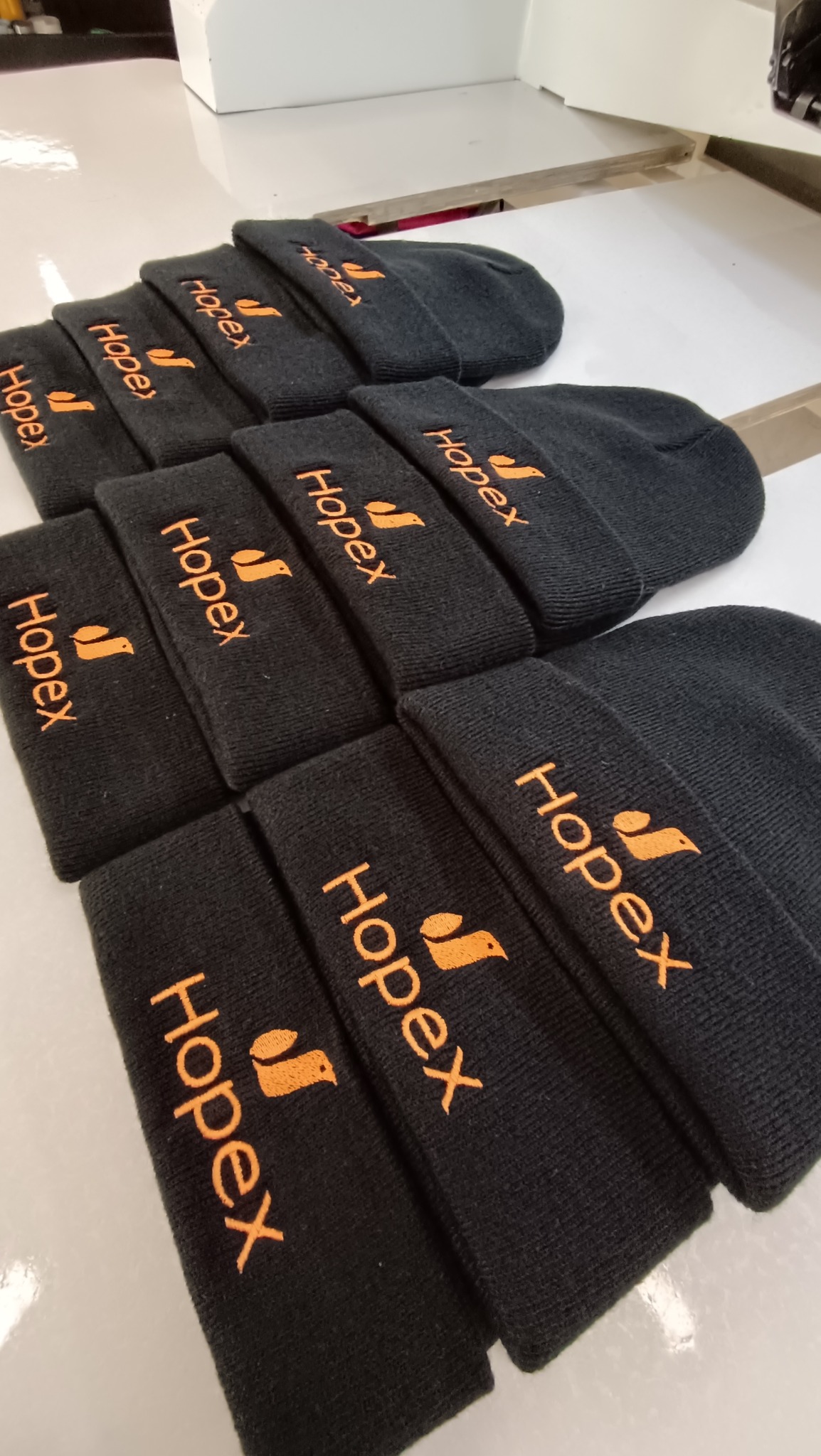 Czarne czapki zimowe z wyhaftowanym pomarańczowym logo 'Hopex' ułożone rzędami na białym stole.