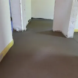 Świeżo wylana, szara posadzka jastrychowa w trakcie schnięcia, widoczne żółte obrzeża przy ścianach i surowy stan wykończenia wnętrza.