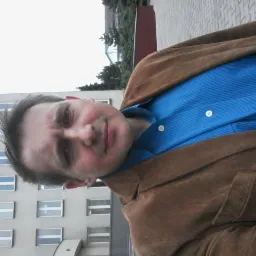 Portret mężczyzny w brązowej kurtce i niebieskiej koszuli na tle budynku z oknami, obrócony kadr.