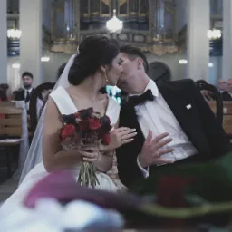 Pocałunek nowożeńców w kościele podczas ceremonii ślubnej, panna młoda trzyma bukiet czerwonych róż, w tle goście siedzący na ławkach.