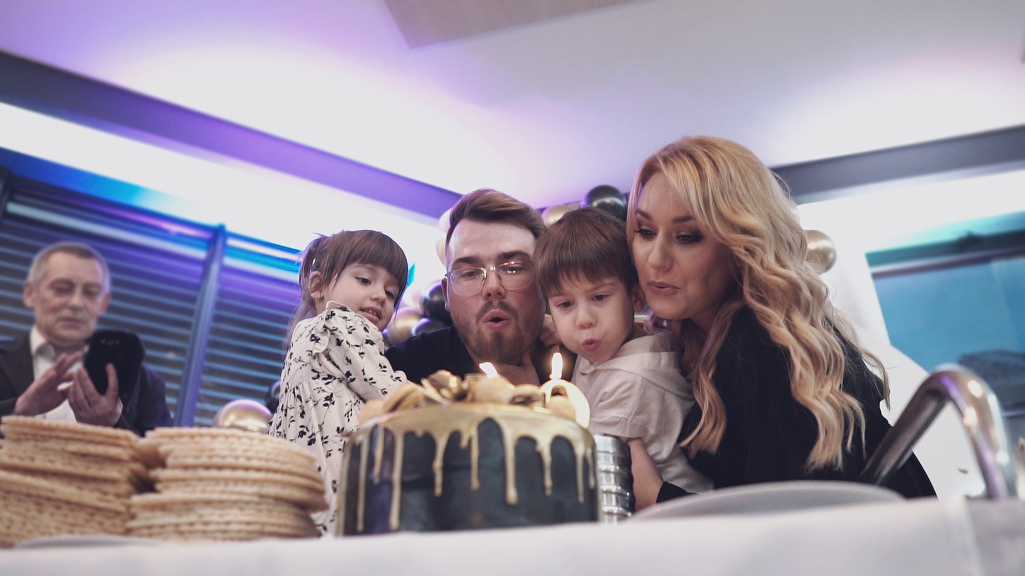 Rodzina z dwójką dzieci zdmuchuje świeczki na czarnym torcie urodzinowym z dekoracją ze złota, w tle mężczyzna robi zdjęcie telefonem.