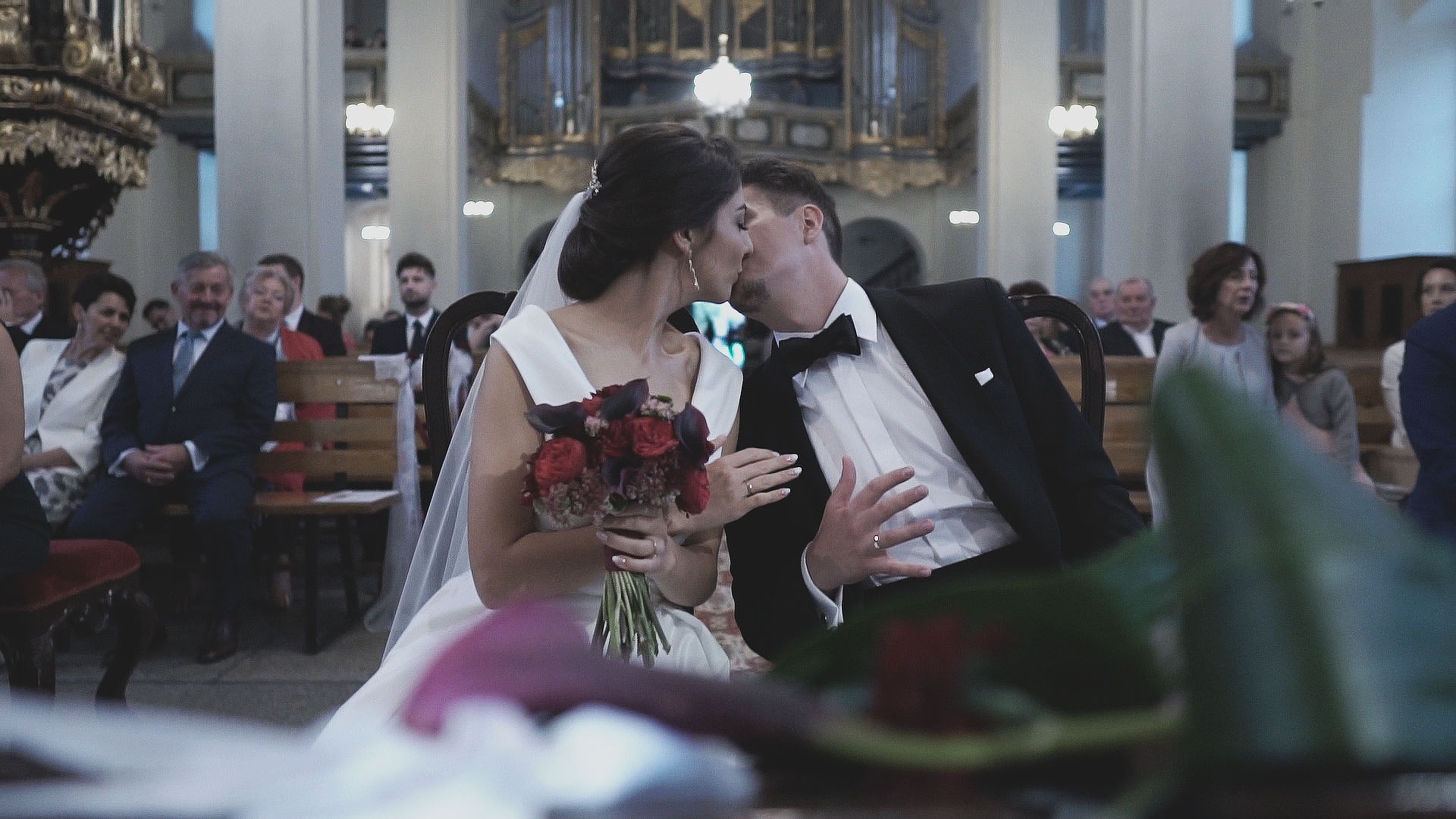 Pocałunek nowożeńców w kościele podczas ceremonii ślubnej, panna młoda trzyma bukiet czerwonych róż, w tle goście siedzący na ławkach.