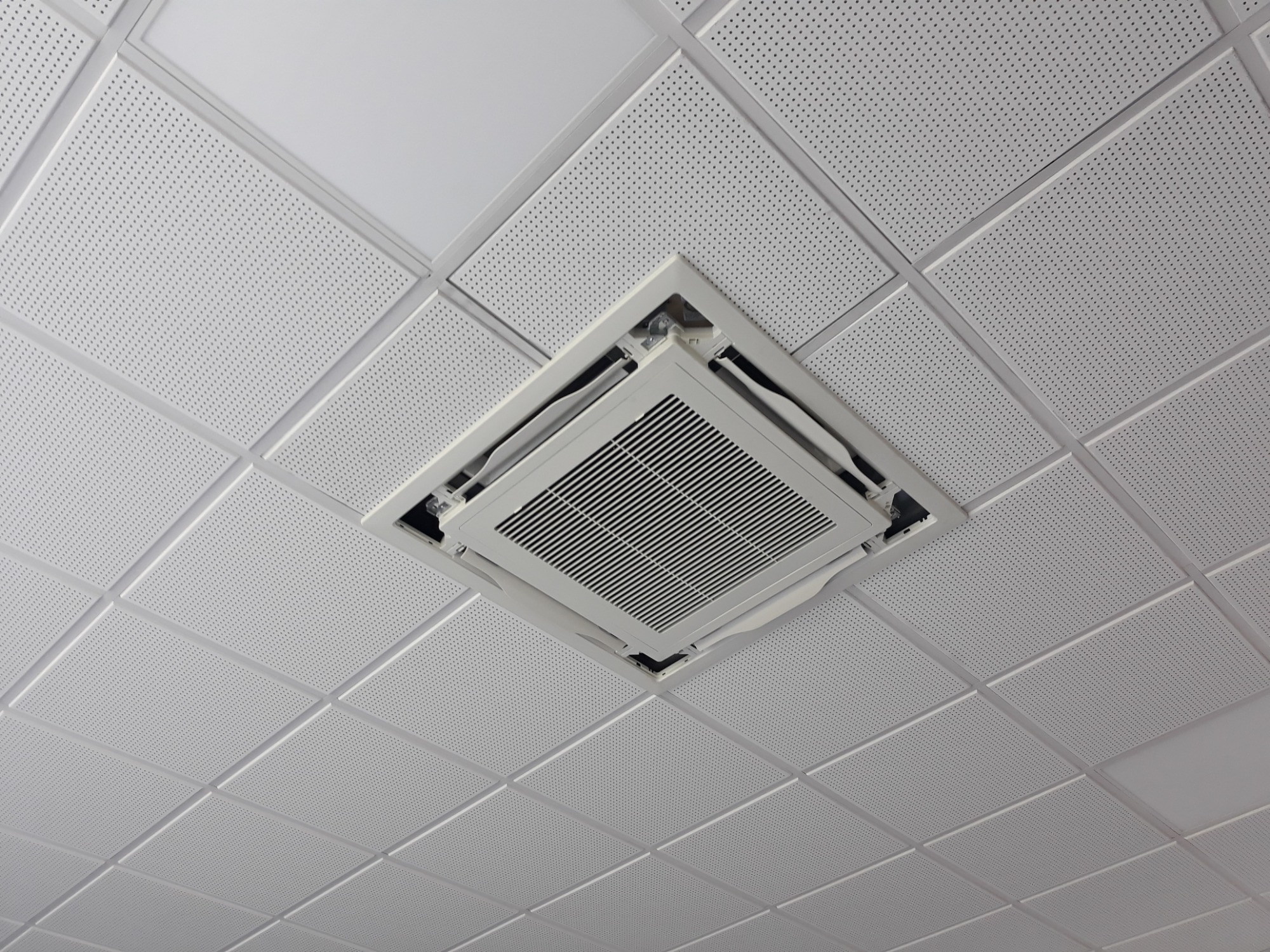 Widok z dołu na kasetonowy sufit z wbudowaną, białą jednostką klimatyzacyjną, widoczne perforowane płyty sufitowe.