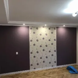 Pokój po odświeżeniu ścian: dwie ściany w kolorze ciemnej śliwki, pomiędzy nimi pas tapety w kwiaty, parkiet w jodełkę, biały sufit z listwami dekoracyjnymi.