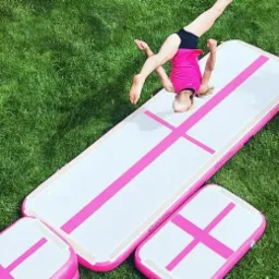 Dziewczynka w różowej koszulce wykonuje salto na białym materacu gimnastycznym z różowymi paskami na trawie.