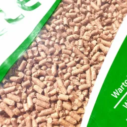 Szczelnie zapakowany worek z pelletem drzewnym, widoczne detale struktury pelletu przez przezroczyste opakowanie, fragment zielonego nadruku na worku.