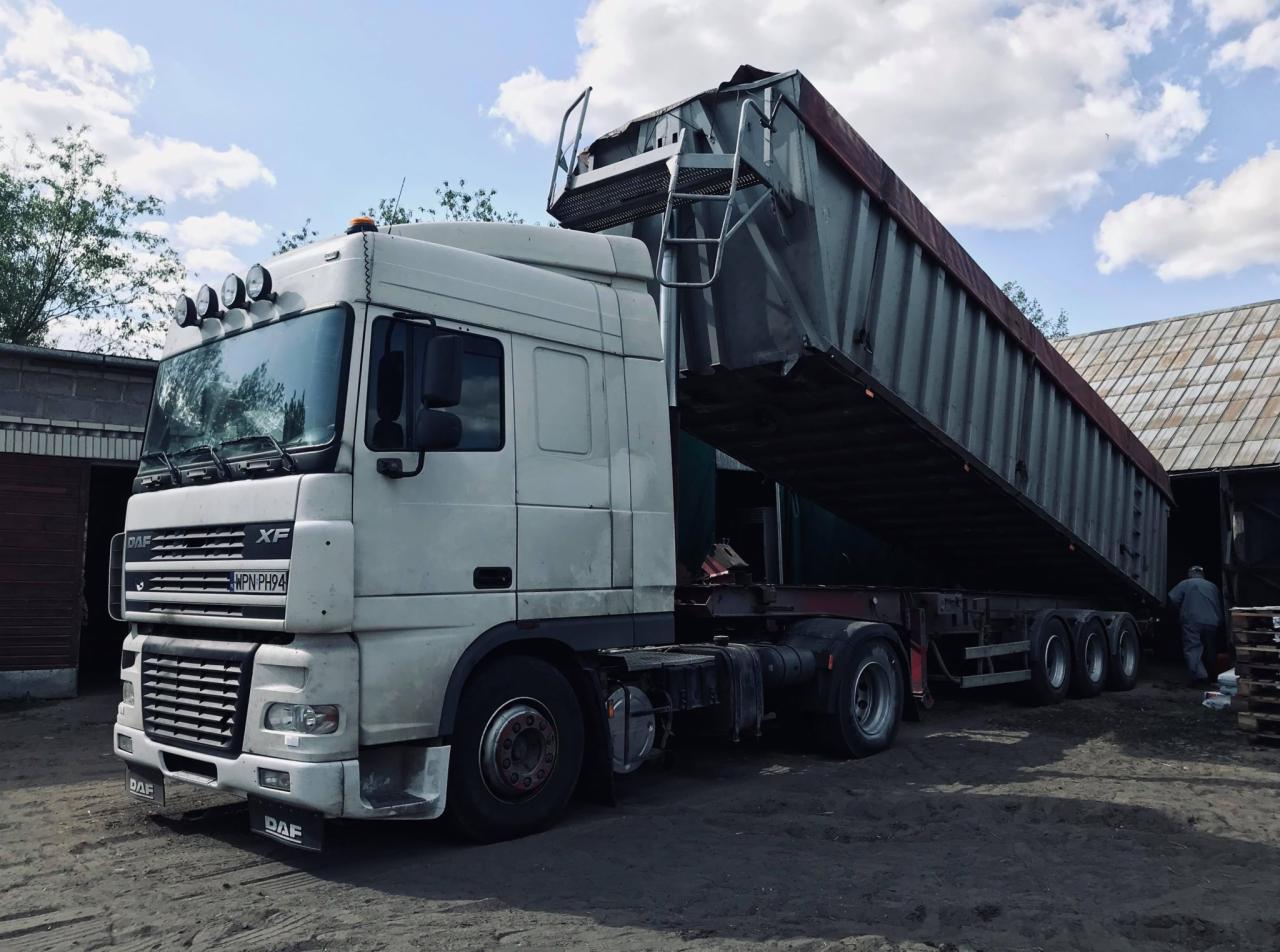 Brudna biała ciężarówka DAF XF z podniesioną naczepą wywrotką, widoczne światła na dachu kabiny, ziemisty plac i budynek w tle.