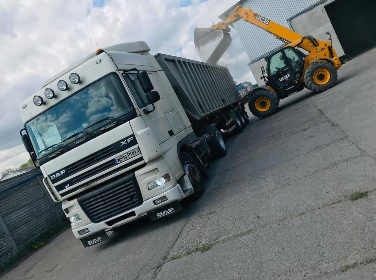 Żółta ładowarka teleskopowa JCB napełnia materiałem sypkim naczepę ciężarówki DAF XF na betonowym placu, widok z dołu.