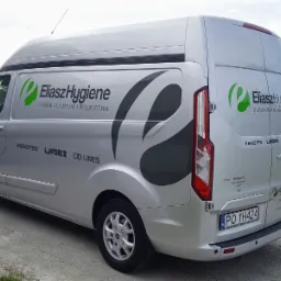 Srebrny samochód dostawczy z logo firmy Eliasz Hygiene i informacjami o chemii dla myjni i rolnictwa, zaparkowany na żwirowej drodze z widokiem na zielone otoczenie.