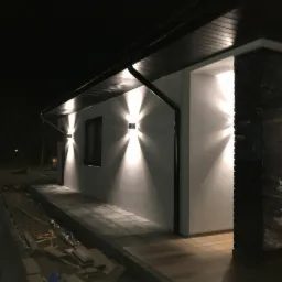 Elewacja budynku z zamontowanym zewnętrznym oświetleniem punktowym, widoczne czarne rynny i kolumna wykończona kamieniem dekoracyjnym, nocne ujęcie.