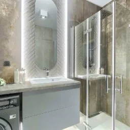 Nowoczesna łazienka z szarymi płytkami imitującymi kamień, owalne lustro z podświetleniem LED, kabina prysznicowa ze szklanymi drzwiami i pralką w zabudowie pod blatem.