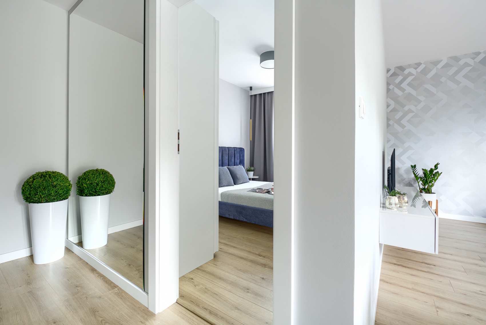 Nowoczesne wnętrze mieszkania z widokiem na sypialnię przez otwarte drzwi, z roślinami w donicach przy lustrzanej szafie i geometryczną tapetą w salonie.