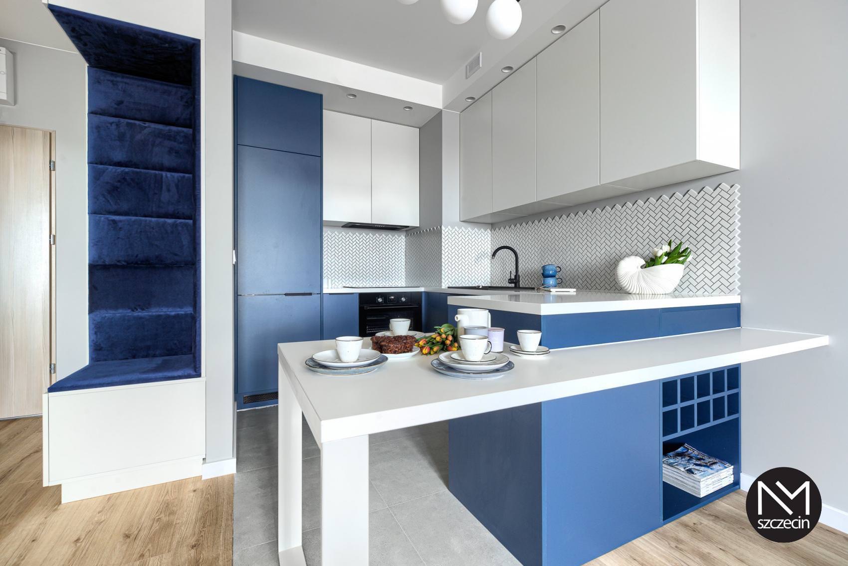 Nowoczesna kuchnia w bieli i odcieniach niebieskiego z wyspą i dekoracyjną ścianą z płytek w jodełkę, uzupełniona o kącik wypoczynkowy z tapicerowanymi panelami ściennymi w kolorze granatowym.