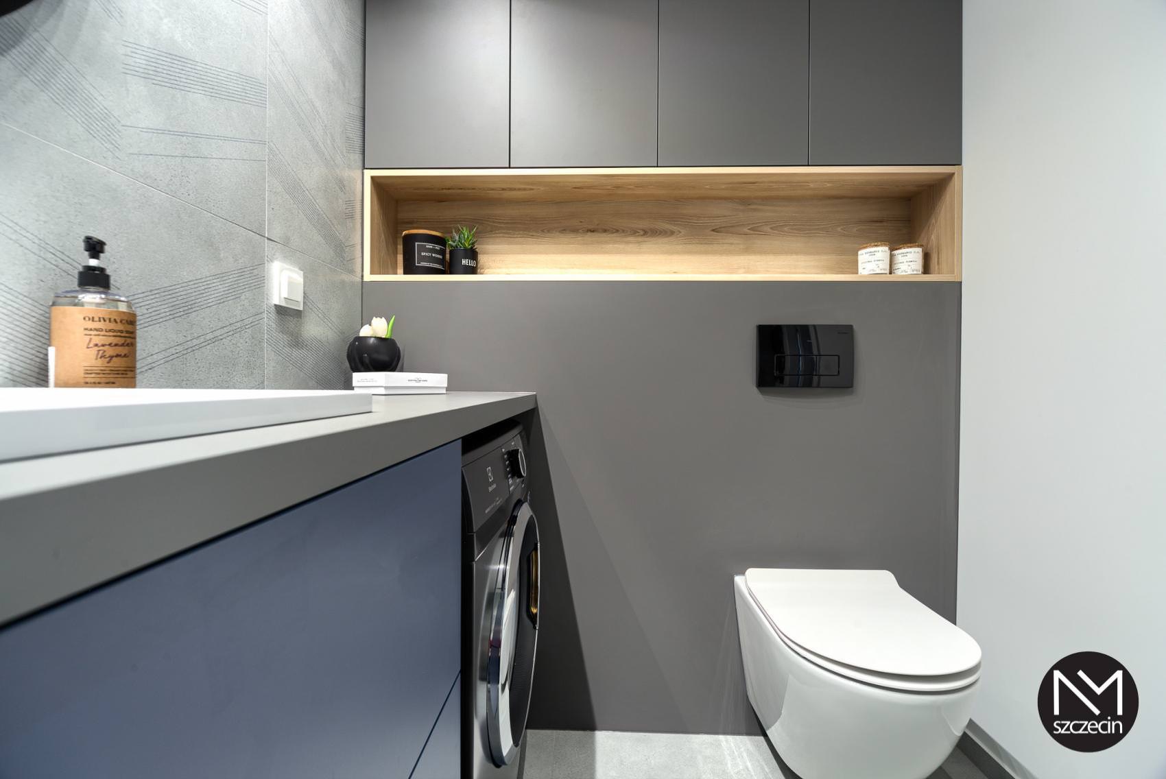Nowoczesna łazienka w Szczecinie z szarymi płytkami, zabudową meblową z drewnianymi akcentami, wiszącą toaletą i pralką w zabudowie pod blatem.